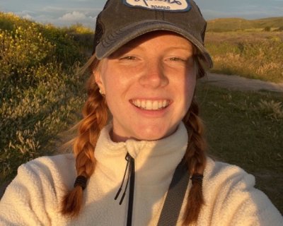 Smiling selfie of Emma at dusk in a field. They wear a baseball cap with a Lucky's logo, their auburn shoulder-length hair is tied into 2 braids.