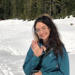 Shadeh waves and smiles at camera, wearing a teal windbreaker, clear glasses, and crossbody purse. They stand on freshly fallen snow with lush pine trees and a clear blue sky in the background. 